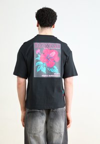 Juodas marškinėliai su dideliu spalvingu hibisko grafiku. Tekstas sako "HAWAII" virš dizaino. Dėvimi su šviesiais pilkais ombre džinsais.
