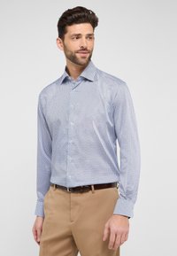 Lichtblauw overhemd met knopen, voorzien van een geometrisch patroon in donkerder blauw, lange mouwen en een klassieke kraag. Geldig met tan kleurige broeken.