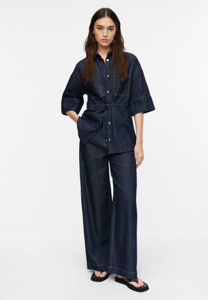 Kobieta ubrana w ciemnoniebieską denimową koszulę ze ściągaczem w talii, szerokie denimowe spodnie i czarne sandały, stojąca na białym tle.