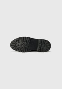 Semelle en caoutchouc noir d'une chaussure, présentant un motif de grip texturé avec des crêtes et des détails circulaires pour une meilleure traction et durabilité.
