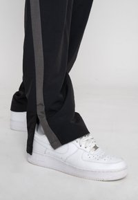 Pantaloni neri con una striscia laterale grigia, caratterizzati da una vestibilità ampia e dettagli con zip vicino all'orlo, abbinati a sneaker bianche in pelle texturizzata.