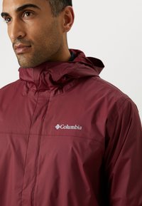 Man draagt een bordeauxrode Columbia regenjas met capuchon, kijkt naar links tegen een effen witte achtergrond.