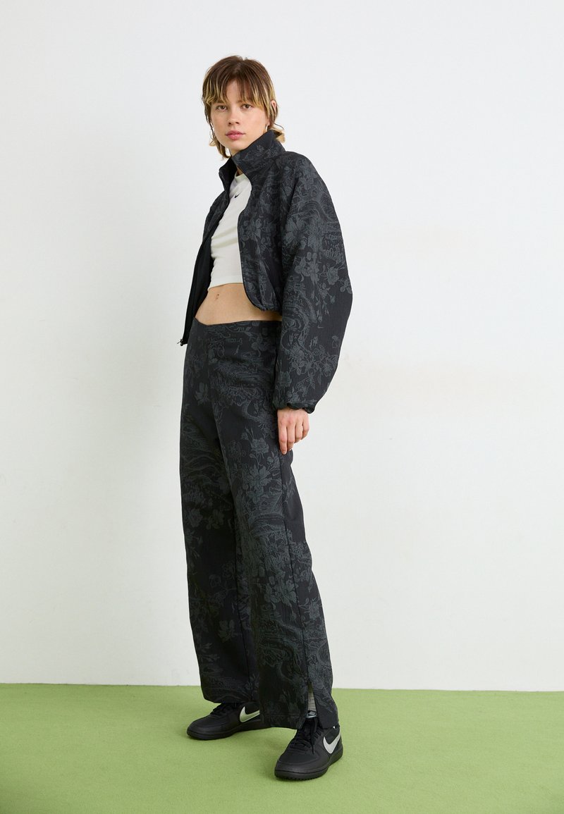 Giacca cropped nera a fantasia floreale abbinata a pantaloni palazzo coordinati, entrambi con una finitura tessurizzata. Sneakers nere completano il look.