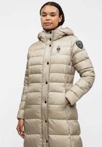 Cappotto piumino beige con cappuccio, caratterizzato da un design trapuntato, bottoni neri e una toppa con logo sulla spalla sinistra.