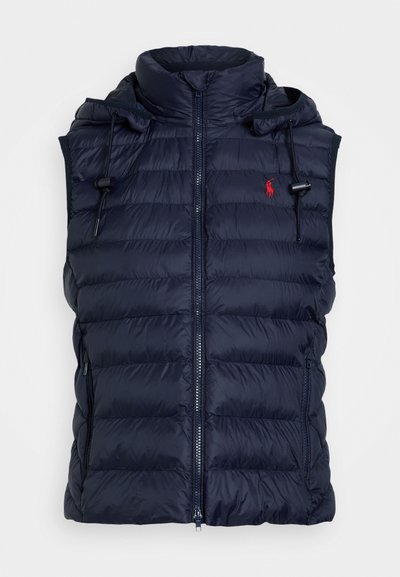 Tumedast sinine puffvest kõrge kraega, lukuga ja reguleeritava nööriga kapuutsiga. Omab horisontaalselt tikitud sektsioone ja küljetaskuid.