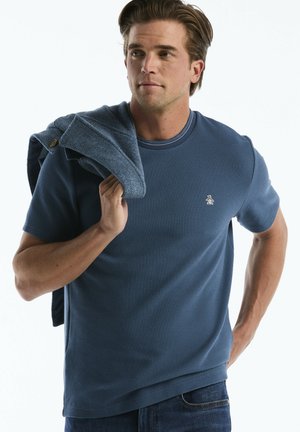 Man in blue textured short sleeve shirt holds a folded blue sweater over his shoulder, looking slightly to his left.