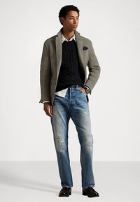 Homme portant un blazer gris, un pull en maille noir, une chemise blanche, un jean bleu déchiré et des chaussures noires, se tenant avec une main dans la poche.