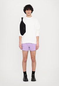 Hvid sweatshirt, lilla shorts og sorte sneakers. En sort crossover taske hænger på den ene skulder. Enkelt design, ingen synlige mønstre.