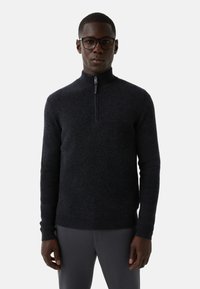 Mężczyzna w czarnej dzierganej bluzie z półsuwnym zamkiem i ciemnoszarych spodniach, stojący ze zrelaksowanymi rękami na tle jasnym.