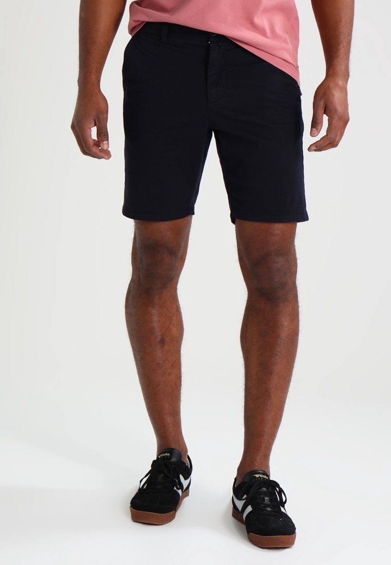 Homme portant un short noir jusqu'aux genoux, des baskets noires et blanches, et une chemise rose, debout devant un fond clair uni.