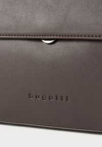 Braune Lederhandtasche mit glatter Textur, ausgestattet mit einem silbernen Druckverschluss und geprägtem "bugatti"-Logo am Boden.