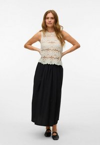 Bílé krajkové top bez rukávů s květinovými vzory a vlnitým lemem, doplněný dlouhou černou sukní. Modelka stojí se s rukama na bocích.