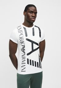 Λευκό κοντομάνικο μπλουζάκι με μαύρο κείμενο και γραφικά στοιχεία. Το υλικό φαίνεται μαλακό, με εφαρμοστή γραμμή και στρογγυλή λαιμόκοψη.