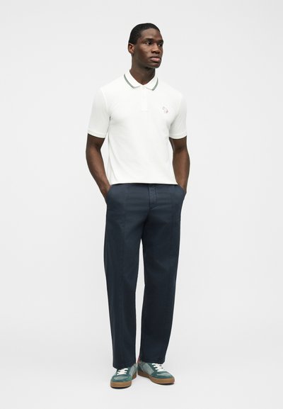 Άνδρας που στέκεται με τα χέρια στις τσέπες, φοράει λευκό πόλο, ναυτικό μπλε παντελόνι και πράσινα αθλητικά παπούτσια μπροστά σε απλό λευκό φόντο.