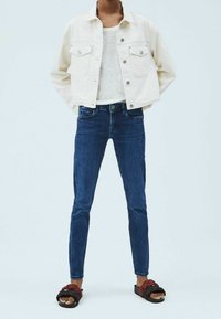 Osoba oblečená v bílém zkráceném saku, průhledném bílém topu, tmavě modrých skinny džínách a černých pantoflích s červenými zapletenými popruhy.