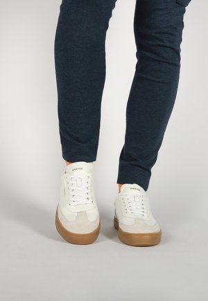 Zapatillas deportivas blancas con detalles de ante, suelas de goma, cordones y marca. Combinadas con pantalones oscuros y ajustados.