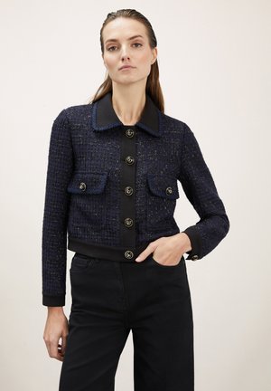 Giacca in tweed blu navy con colletto nero e patta con bottoni, dotata di due tasche sul petto, bottoni dorati e un design corto. Tessuto texture.