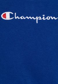 Tejido de algodón azul con una textura suave, que presenta un logotipo bordado en blanco de "Champion" y un acento rojo en la letra "C".