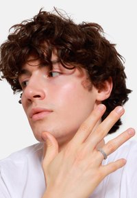 Zilverkleurige ring met een textuur en een geweven patroon, gedragen aan een hand. De model heeft krullend bruin haar en een lichte huid, en draagt een eenvoudig wit shirt.