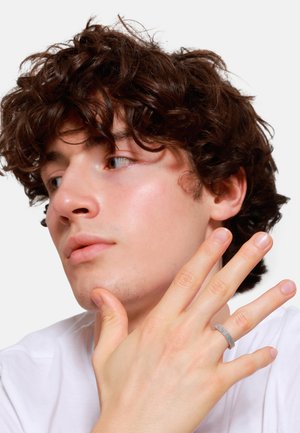 Anello argentato con un motivo tessuto e testurizzato indossato su una mano. Il modello ha capelli ricci castani e pelle chiara, indossa una camicia bianca semplice.