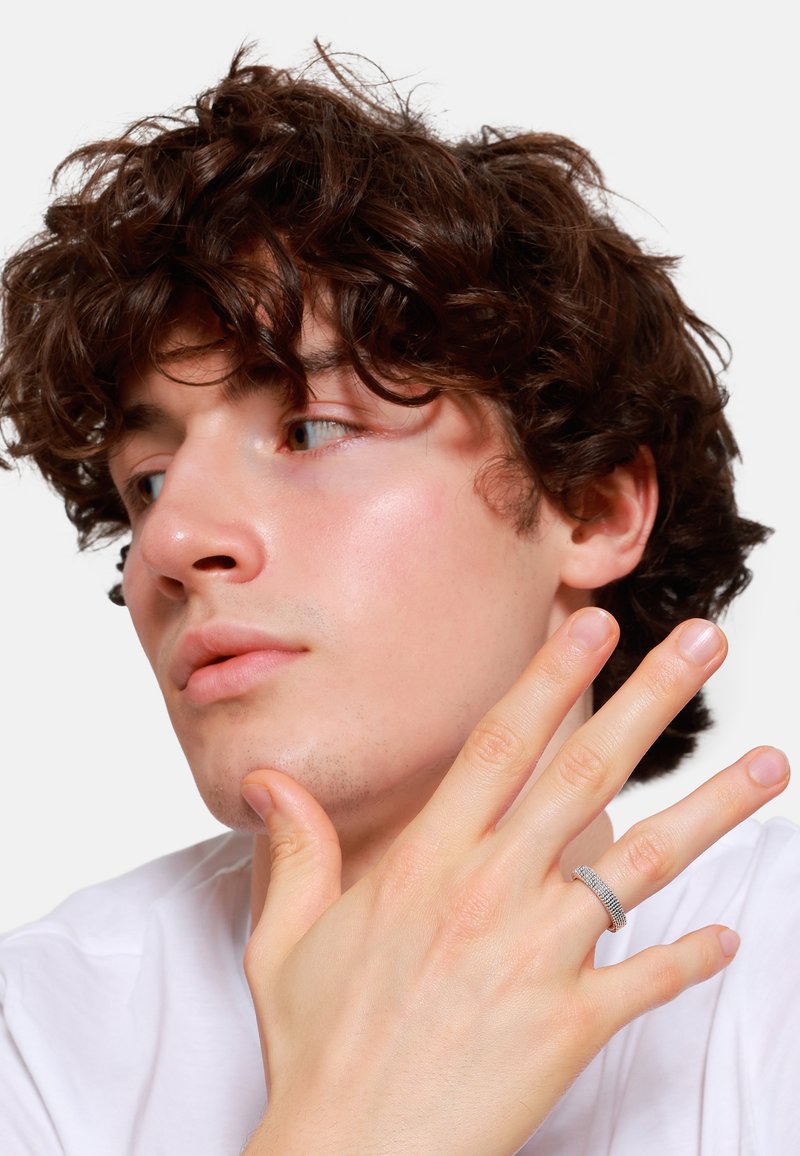 Zilverkleurige ring met een textuur en een geweven patroon, gedragen aan een hand. De model heeft krullend bruin haar en een lichte huid, en draagt een eenvoudig wit shirt.