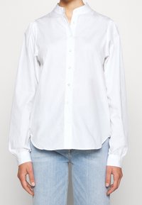 Chemise blanche à boutons avec col montant, manches longues à poignets boutonnés et ourlet arrondi, fabriquée en tissu de coton léger.