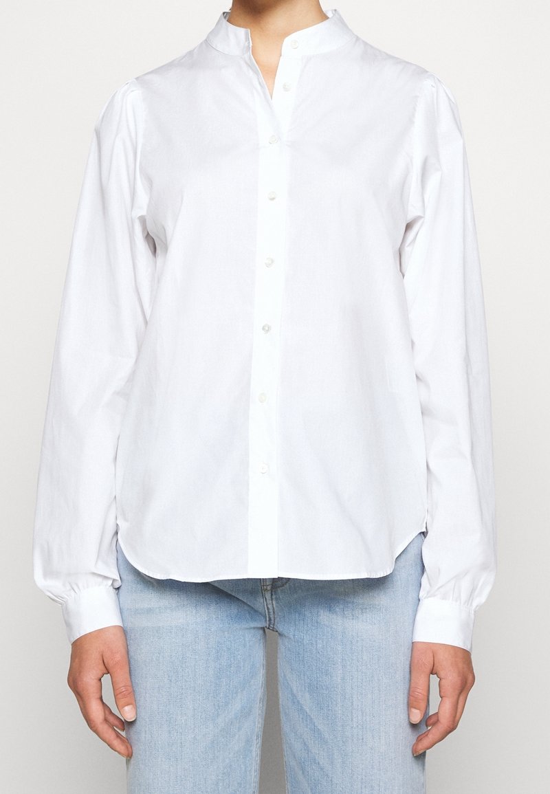 Chemise blanche à boutons avec col montant, manches longues à poignets boutonnés et ourlet arrondi, fabriquée en tissu de coton léger.