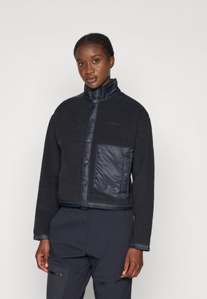 Femme portant une veste polaire noire à boutons-pression et une grande poche en nylon sur le côté gauche, associée à un pantalon foncé.