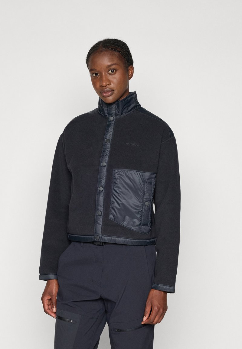 Giacca in pile nera con colletto alto; presenta una tasca in nylon blu contrastante; bottoni a pressione; tessuto testurizzato e particolari lisci.