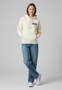 Felpa crema con la scritta "AMOUR" stampata in nero, dotata di tasca frontale e cappuccio con lacci, abbinata a jeans azzurri chiari e sneakers bianche.