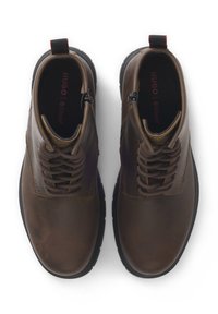Un par de botas de tobillo de cuero marrón con suelas y cordones negros, vistas desde arriba sobre un fondo blanco.