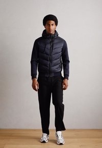Navy puffer jacket with vertical quilted pattern, zip closure, and hood; paired with black trousers and white sneakers. Wood floor background.