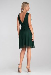 Grünes Kleid mit tiefem V-Rücken, tailliertem Oberteil, flauschigem Tüllrock und auffälligem Krawattenbund. Getragen mit Riemchenschuhen.
