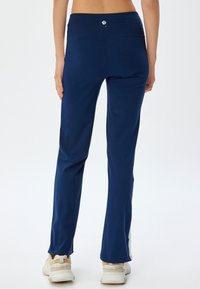 Marineblauwe sportbroek met een hoge taille, gladde textuur en splits aan de enkels, gecombineerd met lichtgekleurde sneakers.