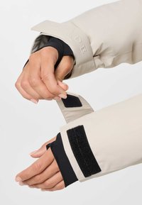 Χέρι ρυθμίζει μαύρο ιμάντα velcro σε μανίκι μπεζ μπουφάν πάνω από μαύρο εσωτερικό στρώμα σε ανοιχτό φόντο.