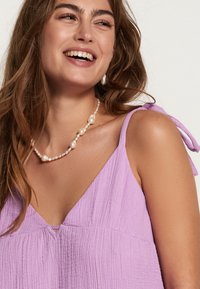 Top texturat în nuanța de liliac, cu decolteu în V și bretele subțiri legate la umeri. O necklace din perlă pune în valoare decolteul. Tesătură ușoară și respirabilă.