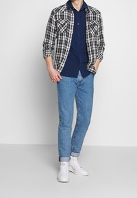 Camicia a quadri bianca e nera indossata sopra una camicia blu navy con bottoni, abbinata a jeans azzurri e sneakers bianche. Vestibilità casual, risvolti dei jeans arrotolati visibili.