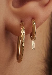 Boucles d'oreilles en or en forme de créole avec une surface texturée et une finition lisse. Une boucle d'oreille présente un pendentif rectangulaire suspendu avec un motif.