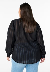 Schwarze, transparente Bluse mit langen Ärmeln und vertikalen Streifen, mit Knopfmanschetten und abgerundetem Saum, kombiniert mit hellblauen Jeans.