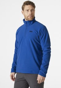 Niebieski polarowy sweter z zamkiem przy kołnierzu, z czarnym logo na piersi. Gładka faktura, długie rękawy i prosta dolna krawędź.