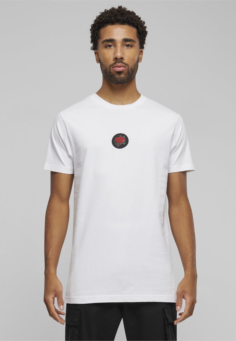 Homme aux cheveux courts et bouclés portant un t-shirt blanc avec un motif de rose rouge sur la poitrine, sur fond gris.