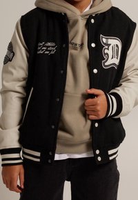 Giacca varsity nera e crema con tessuto testurizzato, polsini a coste e logo ricamato. Indossata sopra un hoodie beige con testo stampato.