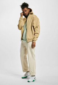 Beige zip-up mikina přes světle zelené tričko, doplněná krémovými kalhotami a bílými teniskami s zelenými akcenty.