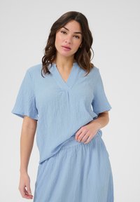 Light blue textured blouse with short sleeves and a V-neck design. Fabric is crinkled, offering a relaxed fit. Matching skirt visible.