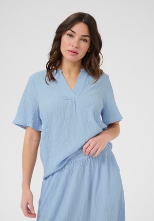 Femme aux cheveux bruns ondulés portant un haut texturé bleu clair à manches courtes et une jupe assortie, debout devant un fond uni.