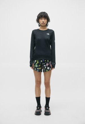 Personne debout portant une chemise noire à manches longues, un short à motifs floraux, des chaussettes noires et des chaussures de trail sur un fond uni.