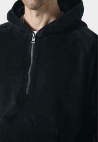 Felpa nera in corduroy con zip frontal, tasca a marsupio e tessuto testurizzato. Il design include un cappuccio aderente e dettagli a coste.