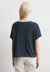 Marineblaues kurzärmliges Hemd aus weichem Stoff. Hat eine entspannte Silhouette und einen abgerundeten Halsausschnitt. Wird mit hellen Hosen getragen.