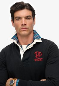 Zwart rugbyshirt met een witte kraag, voorzien van rode borduursels. De binnenkant van de kraag is geruit in blauw en groen. Model draagt een gouden horloge.