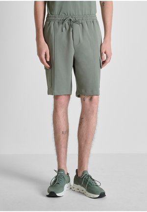 Mann, der grüne Cargo-Shorts mit Kordelzug und passende grüne Sneaker trägt, vor einem schlichten hellgrauen Hintergrund steht.